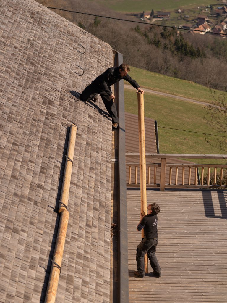 Niels Krüger en équilibre sur un toit, réceptionnant une poutre en bois de la part de Joseph Barotte situé en contrebas